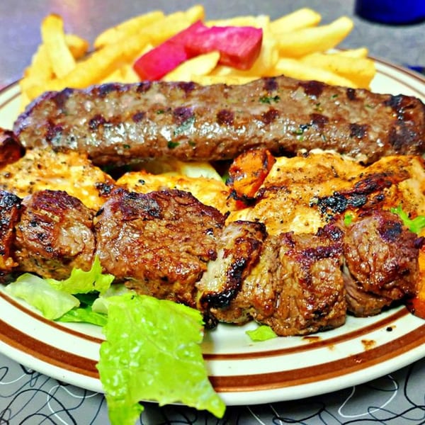 A platter of Mediterranean grilled meats, including a long kabob, chicken, and beef skewers, with fries and pickles.